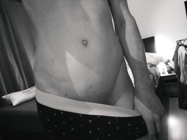 NeueHeimat-hot's bare, slightly chubby belly with a navel, wearing dark, patterned boxer briefs, pulling them down with one hand in a black-and-white photo.