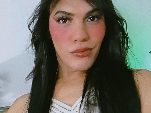 BiancaFerreira has long black hair, pink blush, glossy brown lips, and dark eyebrows. She's wearing a white top with a necklace, looking at the camera with a slight pout.