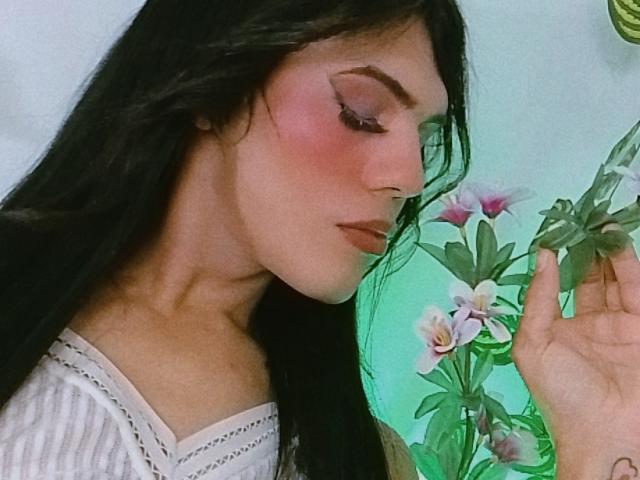 BiancaFerreira with long black hair, pink blush on cheeks, looking down, holding green leaves with white and pink flowers, wearing white off-shoulder top, soft light on face.