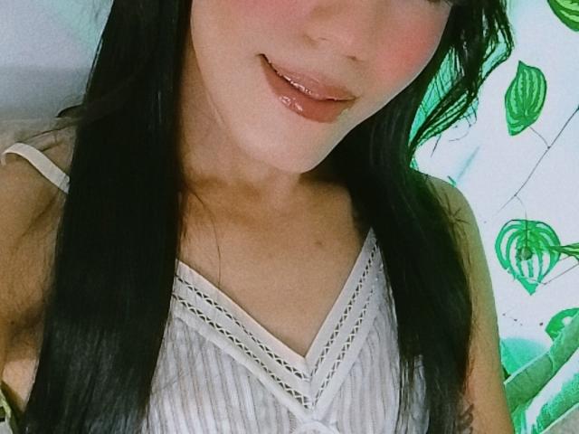 BiancaFerreira's face, showing her shiny pink lips and black hair, wearing a white dress with a V-neck, her small tits slightly visible. Her skin is fair and she's smiling.