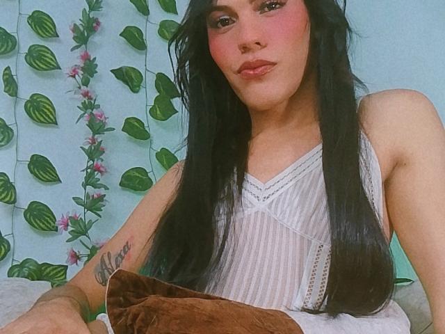BiancaFerreira with long black hair, wearing a white sleeveless dress, holding a brown blanket, has pink blush on cheeks, tattoo on right arm, looking at camera.