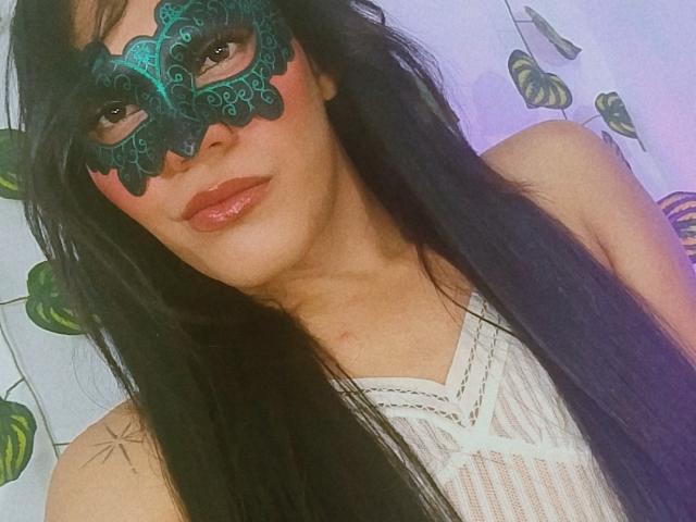 BiancaFerreira with long black hair, wearing a teal lace mask, shiny pink lipstick, and a white sheer top, showing off her tits in the selfie.