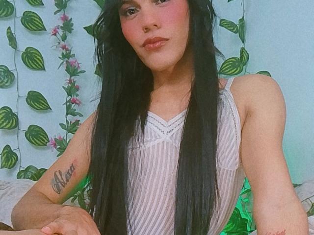 BiancaFerreira with long black hair, wearing a sheer white top, showing a tattoo on her right arm, sitting with a subtle green light in the background.