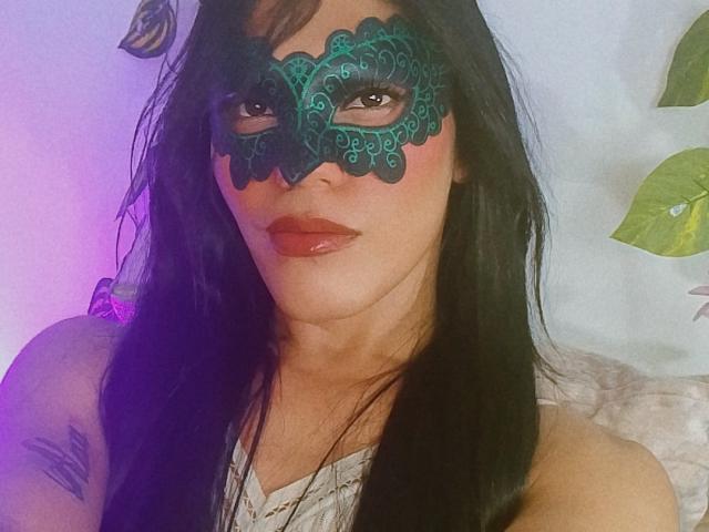 BiancaFerreira with dark green lace mask, red lipstick, and black hair, showing off a tattoo on her right shoulder, sitting on a white pillow. Her pussy is not visible.