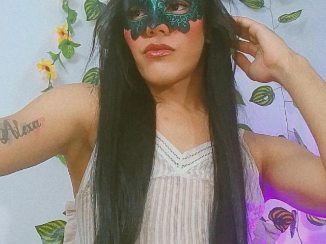 BiancaFerreira with long black hair, wearing a green lace mask, white striped dress, and showing a tattoo on her arm, has her hand in her hair.
