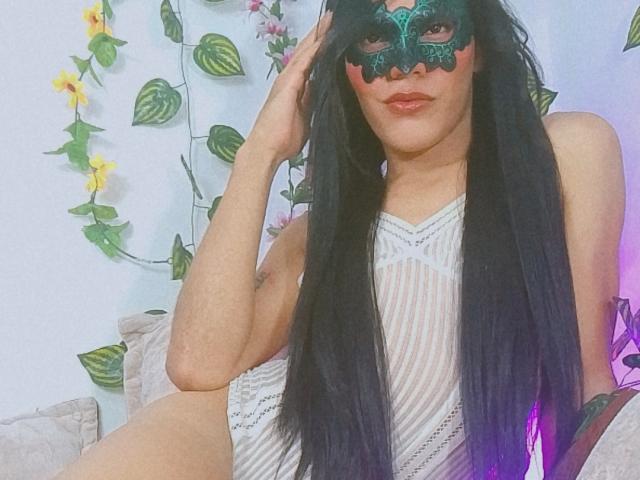 BiancaFerreira with long black hair, green masquerade mask, and white striped dress, sitting with hand on head, showing her ass and soft skin, on a couch.