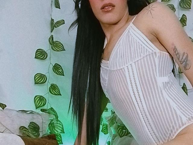 BiancaFerreira with long black hair, wearing a white, sheer, striped, sleeveless top, showing her small tits, tattoo on left arm, green leafy vines in background, soft green light.