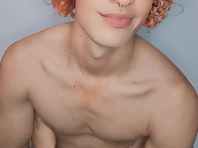 BellaDollI has pale skin, pink curly hair, a nose ring, and is topless with visible collarbones and small moles on her chest. Her pink lips are slightly parted.