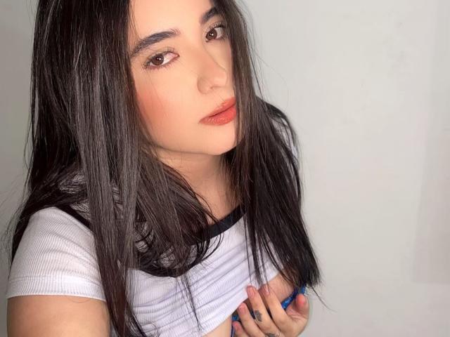 LianaBernocchi with long black hair, brown eyes, and red lips, wearing a white crop top, holding her tits, blue nails, and a small tattoo on her left hand.