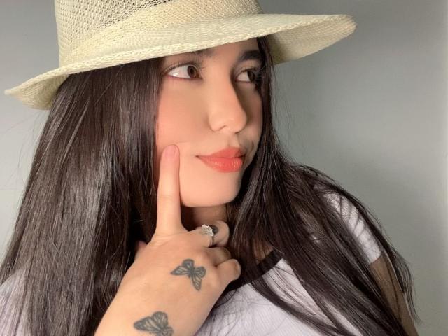 LianaBernocchi with long dark hair, wearing a beige hat, touching her chin with a finger, showing butterfly tattoos on her hand, smiling with pink lipstick.