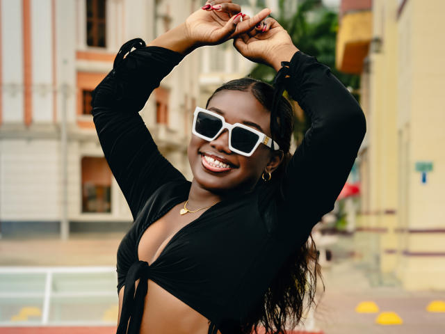 KarlieMoss with dark skin, smiling, wearing white sunglasses, black tied-up top, showing tits, arms raised, red nails, gold necklace, black hair, and gold hoop earrings.