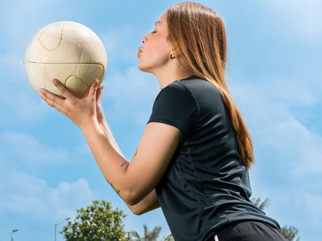 VictoriaOrtega with long brown hair, black t-shirt, and black pants, holding a white soccer ball, kissing it, against a blue sky. Her tits are slightly visible under the shirt.