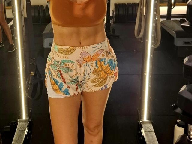 AnneMilfCharm with tan skin, small tits in an orange sports bra, and colorful floral booty shorts, standing on a black gym machine. Her abs are visible.