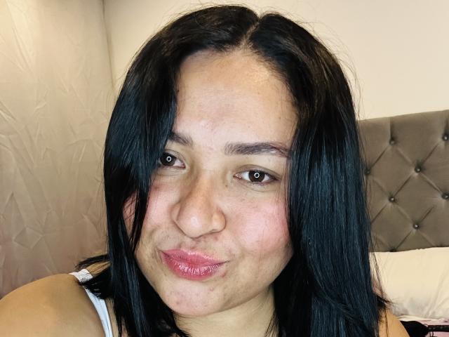 KarlaWild with black hair, brown eyes, and pink lips, pouting her small, slightly upturned nose. Her white tank top is visible. Her ass is not in the image.