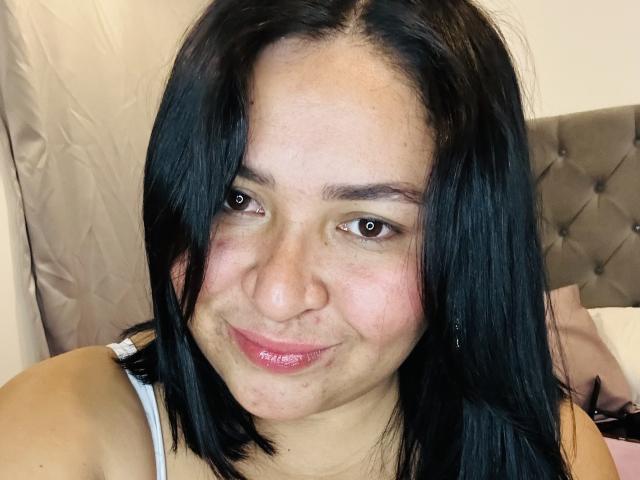 KarlaWild has medium brown skin, black straight hair, brown eyes, and a subtle smile with pink glossy lips. She's wearing a white tank top, showing her small tits.
