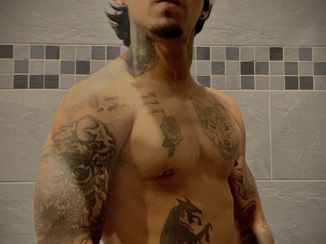 Rv69energy, shirtless with tattoos covering arms and chest, including a rose and skull, has a muscular build and short dark hair, standing in a tiled bathroom.