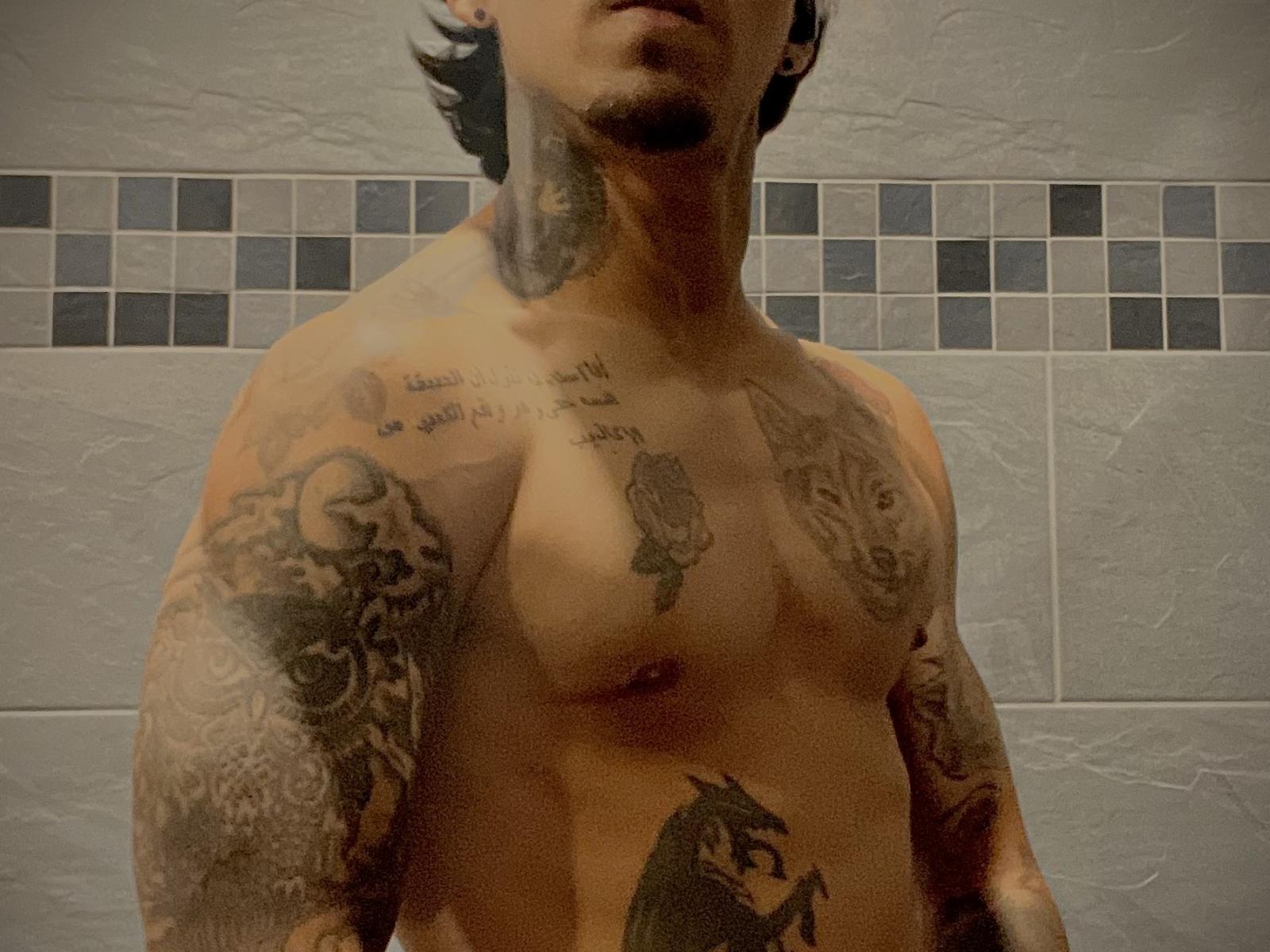 Rv69energy, shirtless with tattoos covering arms and chest, including a rose and skull, has a muscular build and short dark hair, standing in a tiled bathroom.