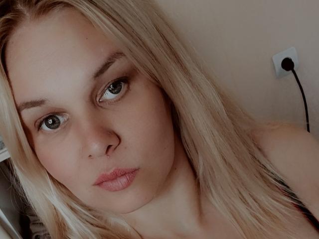 LeilaBlossom has blonde hair, green eyes, and pouty pink lips. She's looking straight at the camera with a slightly bored expression. Her nose is small and her skin is fair.