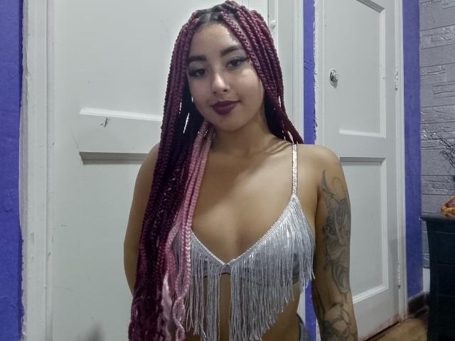 AryVixen with pink and purple braids, wearing a silver fringe bikini top, showing her tits and tattoo on her right arm, smiling with dark lipstick.