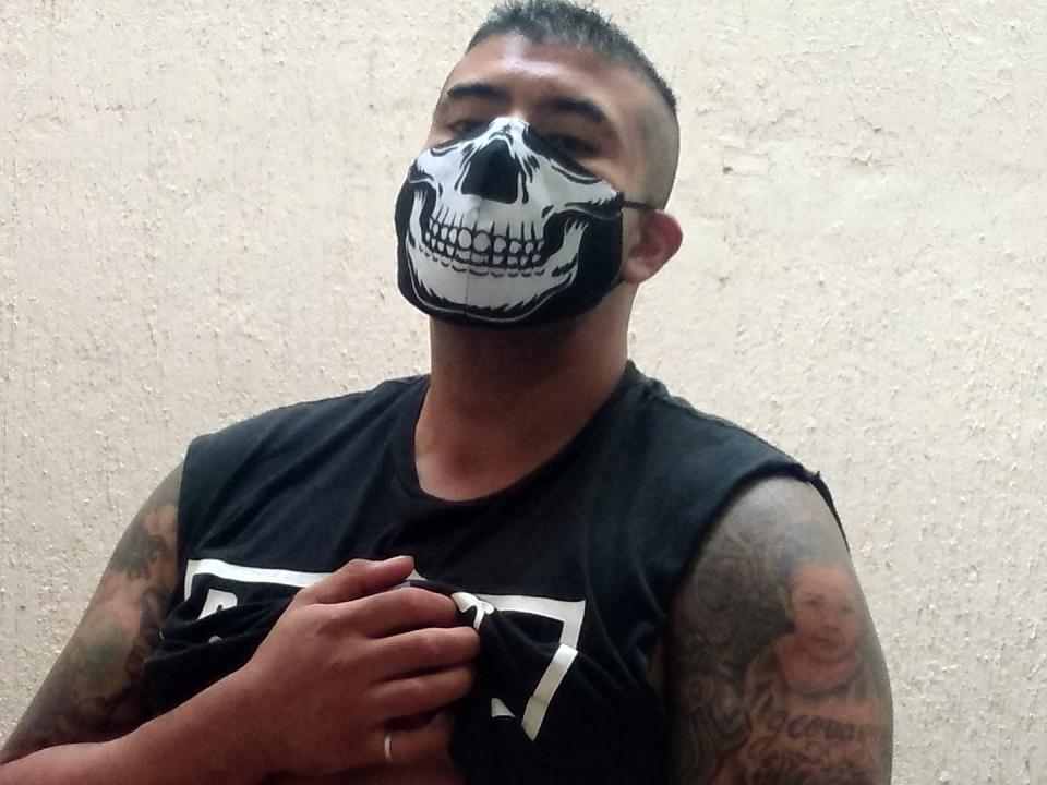 CholoBlakI with a skull mask, black sleeveless shirt, tattoos on arms, short buzzcut, holding shirt, textured beige wall in background.