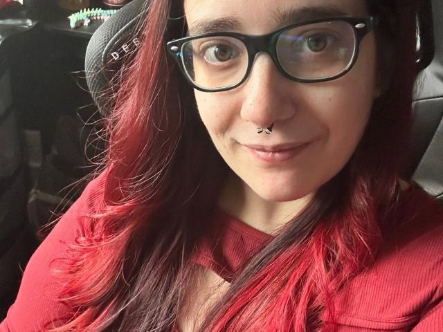 LadyJosefine-hot with red-dyed hair, black glasses, nose piercing, and a subtle smile, wearing a red shirt, seated in a car, looking at the camera.