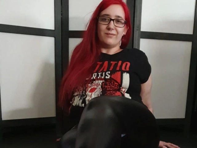 LadyJosefine-hot with red hair, glasses, and nose ring, wearing a black t-shirt and black pantyhose, sitting with her legs crossed, showing her black pantyhose.