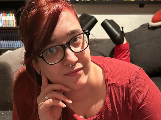 LadyJosefine-hot with red hair, glasses, nose ring, and red shirt, resting on a couch, legs up, wearing black high heels, smiling at the camera.
