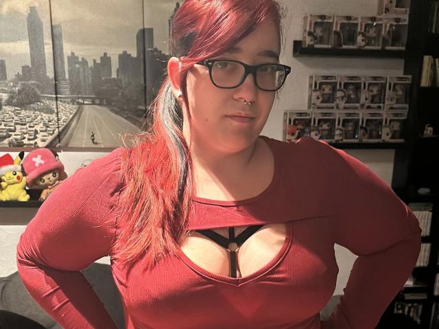 LadyJosefine-hot with red hair, glasses, nose ring, wearing a red long-sleeve top with a cleavage cutout, showing her tits, standing in a sunlit room.