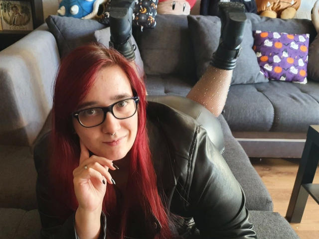 LadyJosefine-hot with red hair, black glasses, leather jacket, and black boots, lying on a gray sofa, legs up, showing her ass, smiling, and touching her chin.