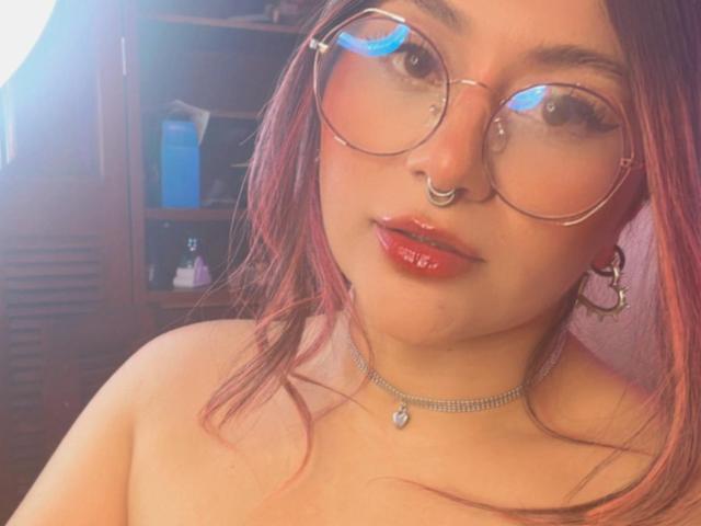 LorraineV has red lipstick, nose ring, round glasses, and pink-dyed hair, wearing a necklace with a small pendant, showing bare shoulders. Her tits are not visible.