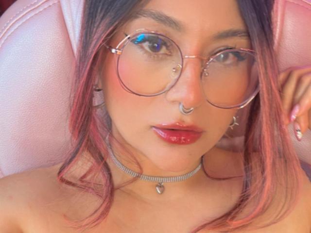 LorraineV with pinkish-red hair, round glasses, nose ring, shiny lips, pink chair, sparkling choker, and pink nails, looking straight at the camera, showing her cute face.