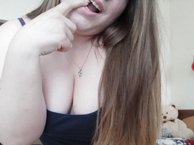 Leiliia with long brown hair, biting her finger, showing off her big tits in a black strap top, wearing a necklace, with a cute teddy bear in the background.