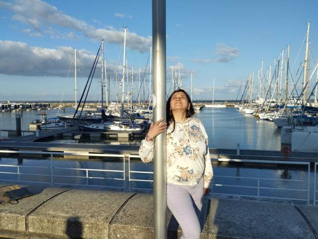 BlissSophia leaning against a metal pole, wearing a floral shirt and purple pants, with her ass slightly pressed against the pole, in front of a marina with boats.