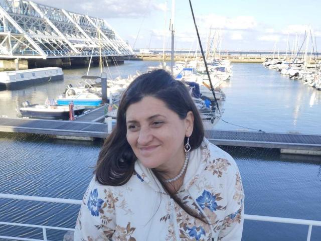 BlissSophia has medium tits, brown hair, and a floral shirt, smiling with a pearl necklace, standing by a marina with boats and a bridge in the background.