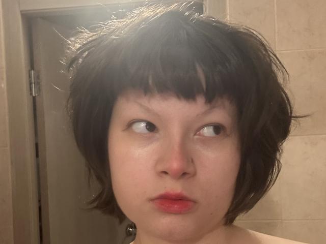 HyldaShirl ha i capelli neri, corti e disordinati, la pelle pallida e il rossetto rosso, con un'espressione accigliata. Sta guardando di lato con un'aria leggermente confusa o infastidita..