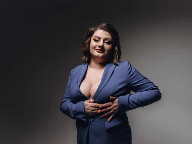 ZannaDreil has big tits, wearing a blue suit, unbuttoned to show her cleavage, with hands on her chest, smiling, brown wavy hair, soft light, gray background.