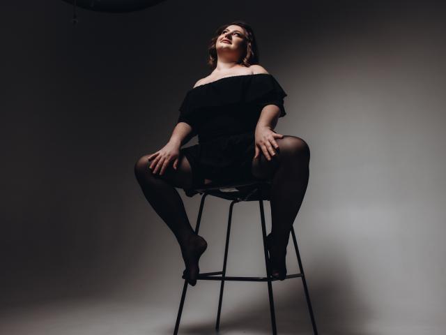 ZannaDreil sits on a stool, legs spread, wearing an off-shoulder black top, black garter stockings, and heels, hands on knees, looking up, with a confident, sexy expression.