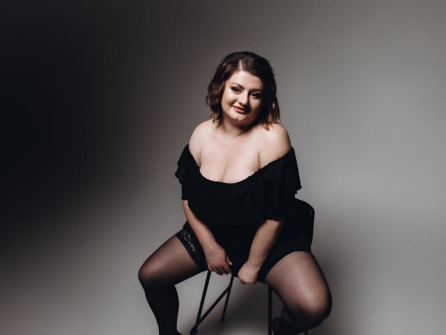 ZannaDreil with wavy brown hair, smiling, sitting on a chair, legs apart, black off-shoulder top, lace stockings, hands on knees, revealing her tits and black lace panties.