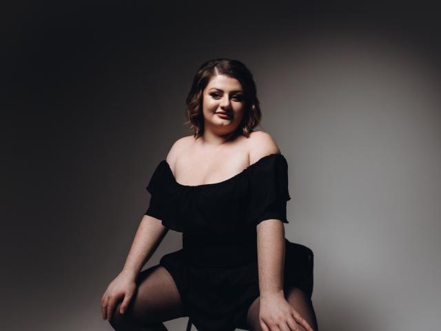 ZannaDreil with shoulder-length brown hair, off-shoulder black dress, sitting with legs apart, showing tits, slight smile, dim lighting, gray background, soft shadows, confident, sexy.