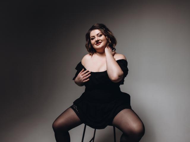 ZannaDreil with curvy body, medium tits, dark brown wavy hair, black off-shoulder dress, black lace stockings, smiling, hand on neck, sitting on chair, legs spread.