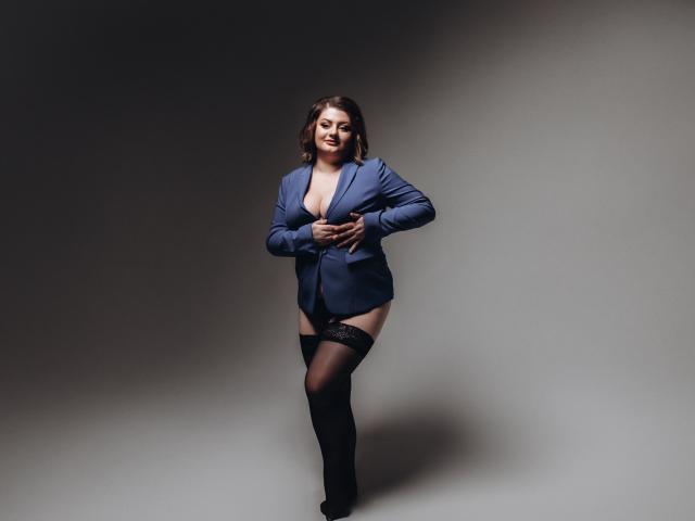ZannaDreil, with dark brown hair, wearing an unbuttoned blue jacket, black lace thigh-highs, and no pants, stands smiling, hands near her chest, exposing her tits.