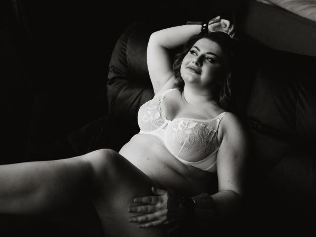ZannaDreil, lying on a dark chair, wearing a white lace bra, hand on belly, legs spread, smiling, black-and-white, soft lighting, relaxed, confident, curvy, busty.