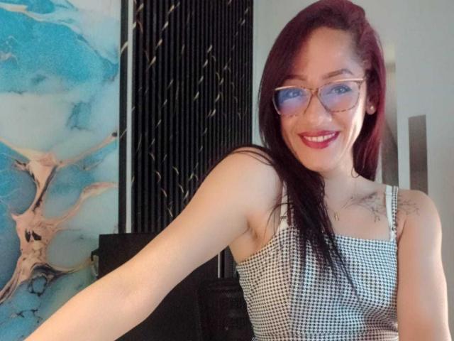 SamanthaCorine, met rood haar, een bril en een glimlach, draagt een geruit top en laat op de selfie haar borsten zien. Haar arm is uitgestrekt, ze maakt de foto zelf..