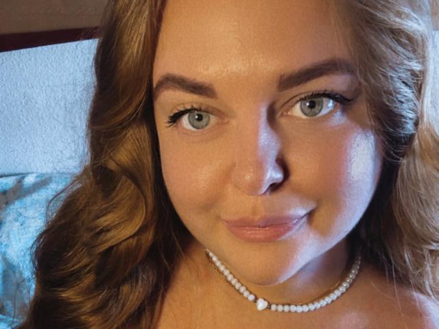 AmeliyaXq has blue eyes, light brown wavy hair, a pearl necklace, and is smiling with pink lips. Her nose is small and her eyebrows are neatly shaped.