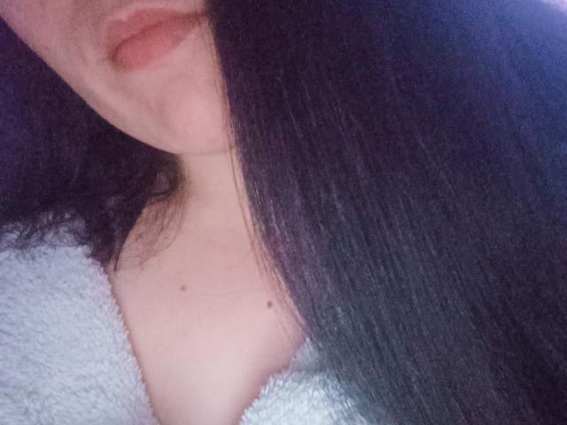 AlikaSun's face with long black hair, slightly parted lips, and a small mole on her chest, wearing a fluffy white robe, showing a bit of her tits.