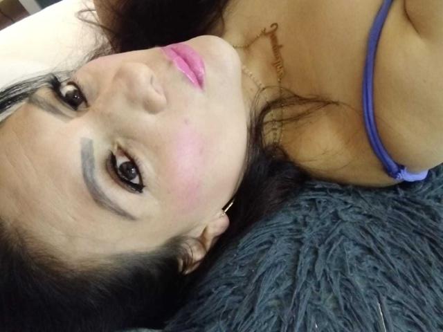 JakyKidman's face with pink lips, dark eyebrows, and a pinkish blush, lying on a gray fuzzy surface, wearing a purple strap, visible breasts, and a gold necklace.