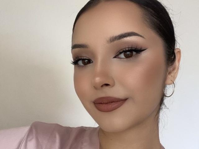 AngelEmma69 has brown eyes, thick eyebrows, long eyelashes, and is wearing pink lipstick. She's looking at the camera with a slight smile, wearing a pink shirt.