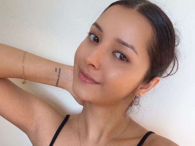 AngelEmma69 with tan skin, brown eyes, and dark hair in a bun, smiling softly, has a small tattoo on her wrist, wearing a black strap top, and a gold bracelet.