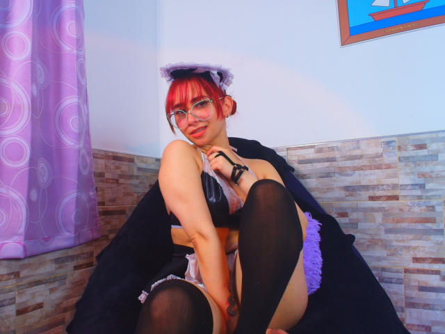 MelannyTaylorr with red hair, glasses, purple cat ears, black bra, purple garter, black stockings, sitting on black cloth, smiling, showing her tits, and a tattoo on her right thigh.