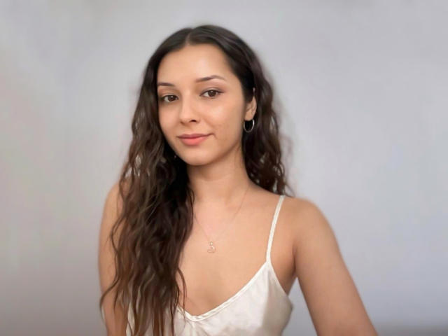 AngelEmma69 with wavy brown hair, light skin, small tits, wearing a white satin camisole, subtle smile, hoop earrings, and a delicate necklace against a plain gray background.